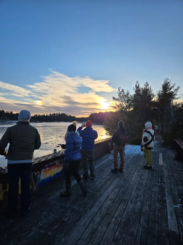 A group of hikers on a trestle bridge overlooking a sunset at Crabtree Neck Land Trust's Old Pond Railway preserve